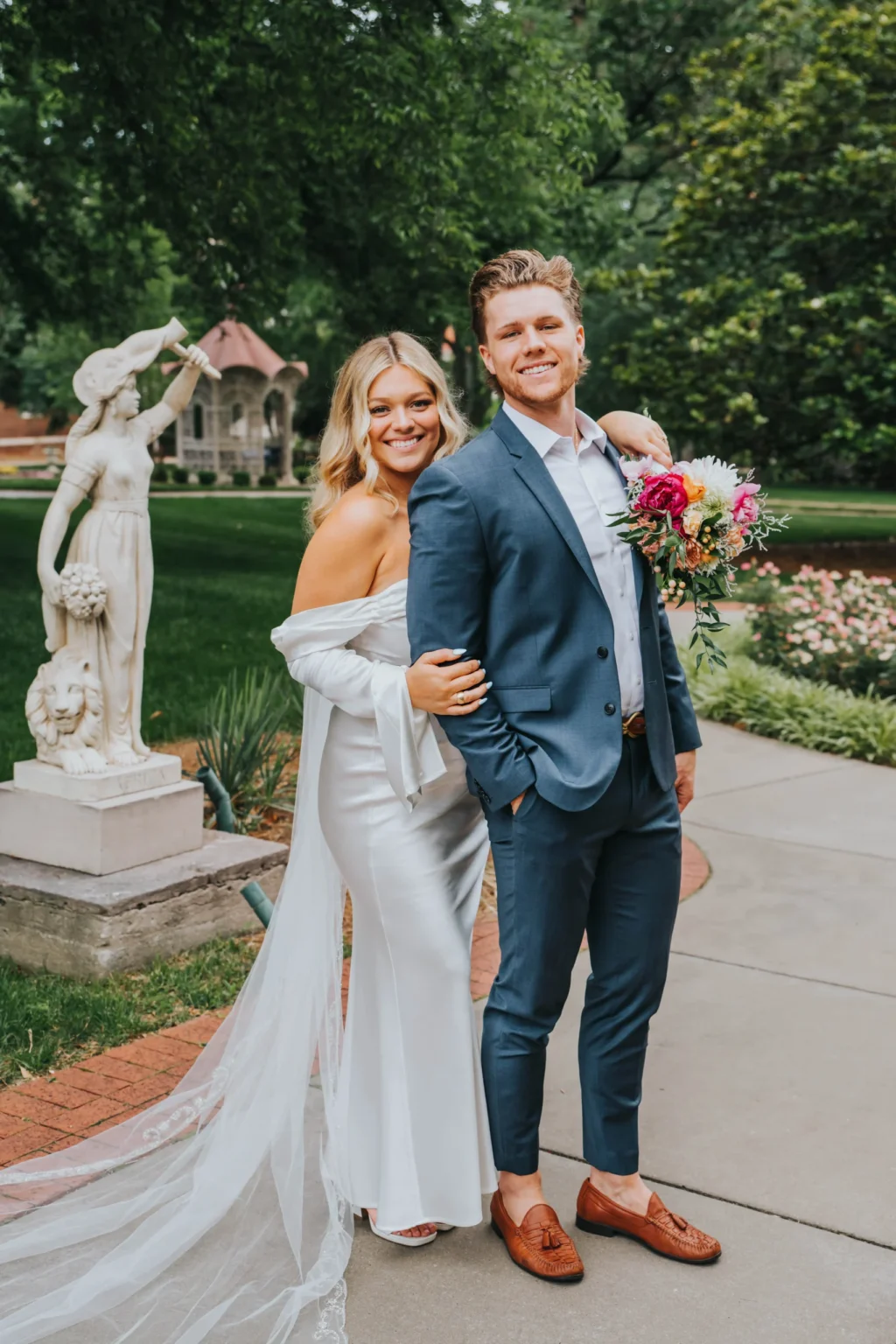 Elopement Wedding A smiling couple stands side by side in a garden. The woman, wearing an off-shoulder white wedding dress with a long veil, has her arm draped over the man's shoulder. The man, in a blue suit and brown shoes, holds a colorful bouquet. A white statue and lush greenery are visible in the background. Elopements Inc