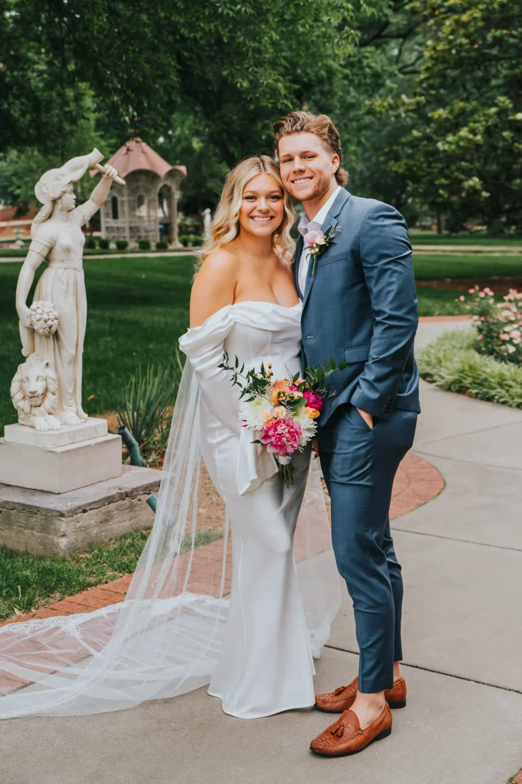 Elopement Wedding A smiling bride in an off-the-shoulder white gown holding a colorful bouquet stands next to a groom in a blue suit and brown shoes. They are posing outdoors, with a statue of a woman holding a basket and a child in the background, and trees and greenery surrounding them. Elopements Inc