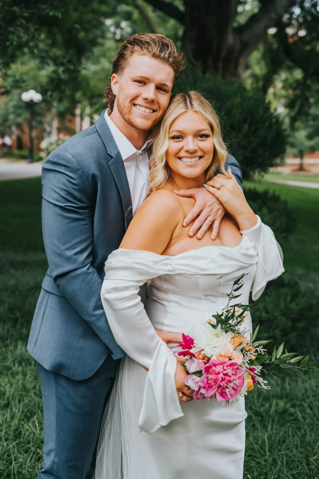 Elopement Wedding A smiling couple poses on a lush lawn with trees in the background. The man, in a blue suit, stands behind a woman in an off-the-shoulder white gown, embracing her. She holds a colorful bouquet of pink, orange, and white flowers. Both appear joyful and relaxed. Elopements Inc