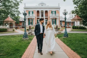 Elopement Wedding A couple dressed elegantly walks hand-in-hand down a brick path towards the camera. The man wears a dark suit, and the woman wears a white dress. They are smiling at each other. Behind them is a grand, old mansion with large pillars, balconies, and ornate lampposts along the pathway. Elopements Inc