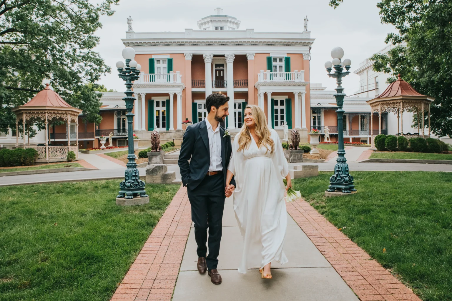 Elopement Wedding A couple walks hand-in-hand down a paved path in front of a grand, historic building with columns and balconies. The man wears a dark suit, while the woman is in a flowing white dress. They both smile warmly at each other. Lush green lawns and two ornate gazebos flank the path. Elopements Inc