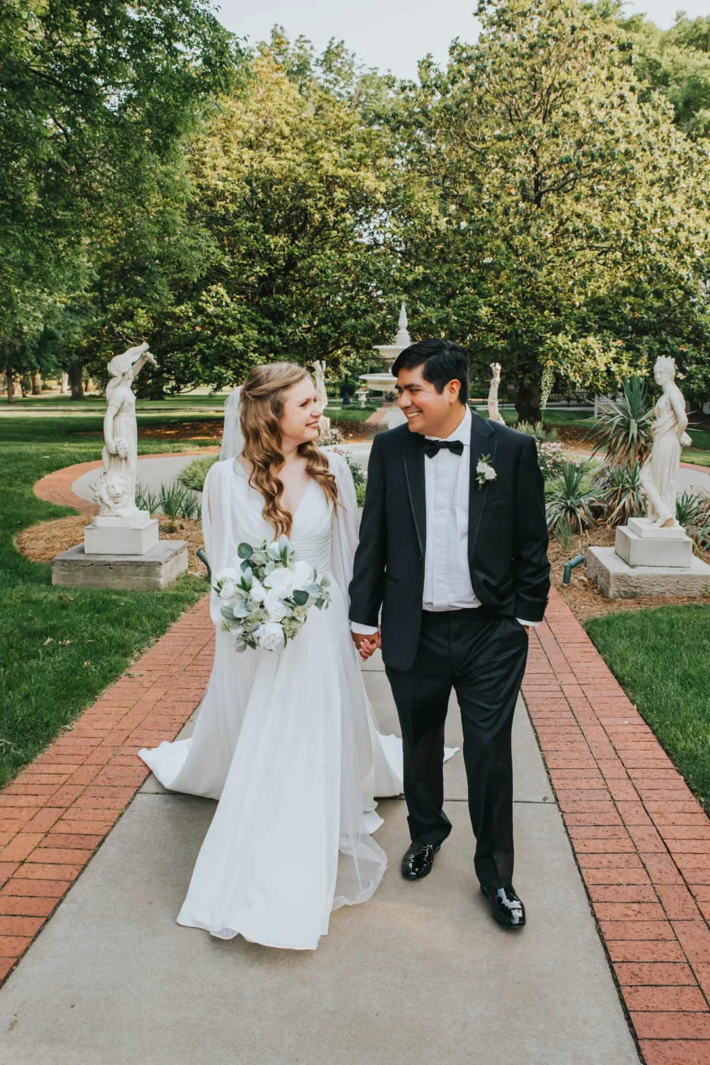 Elopement Wedding A bride and groom walk hand in hand down a brick pathway in a garden. The bride, in a flowing white gown holding a bouquet, smiles at the groom. The groom wears a black tuxedo and bows tie, smiling back at her. Statues and lush greenery surround them on this sunny day. Elopements Inc