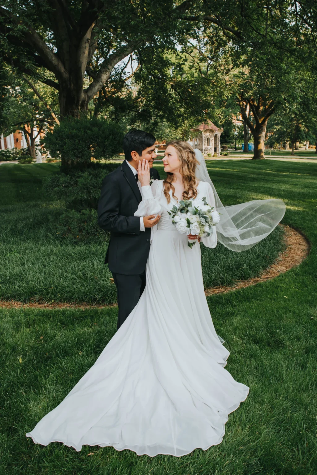 Elopement Wedding A bride and groom stand together outdoors on a lush green lawn surrounded by trees. The bride, in a flowing white gown with a train, holds a bouquet of white flowers and gently touches the groom's face. The groom, in a black suit, tenderly holds the bride's hand. The scene is serene and intimate. Elopements Inc