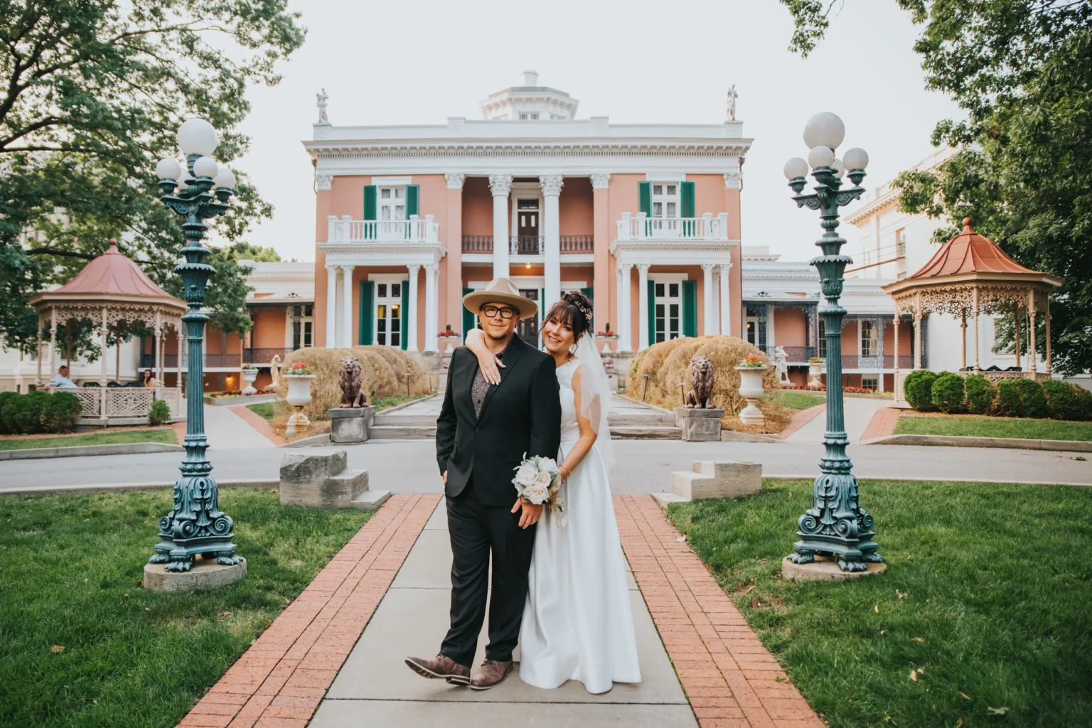 Elopement Wedding A couple in wedding attire poses on a paved pathway leading to a grandiose mansion with a pillared entrance and intricate lamps. The groom, in a black suit and hat, stands beside the bride, who is in a white gown holding a bouquet. Ornate gazebos and lush greenery surround the scene. Elopements Inc