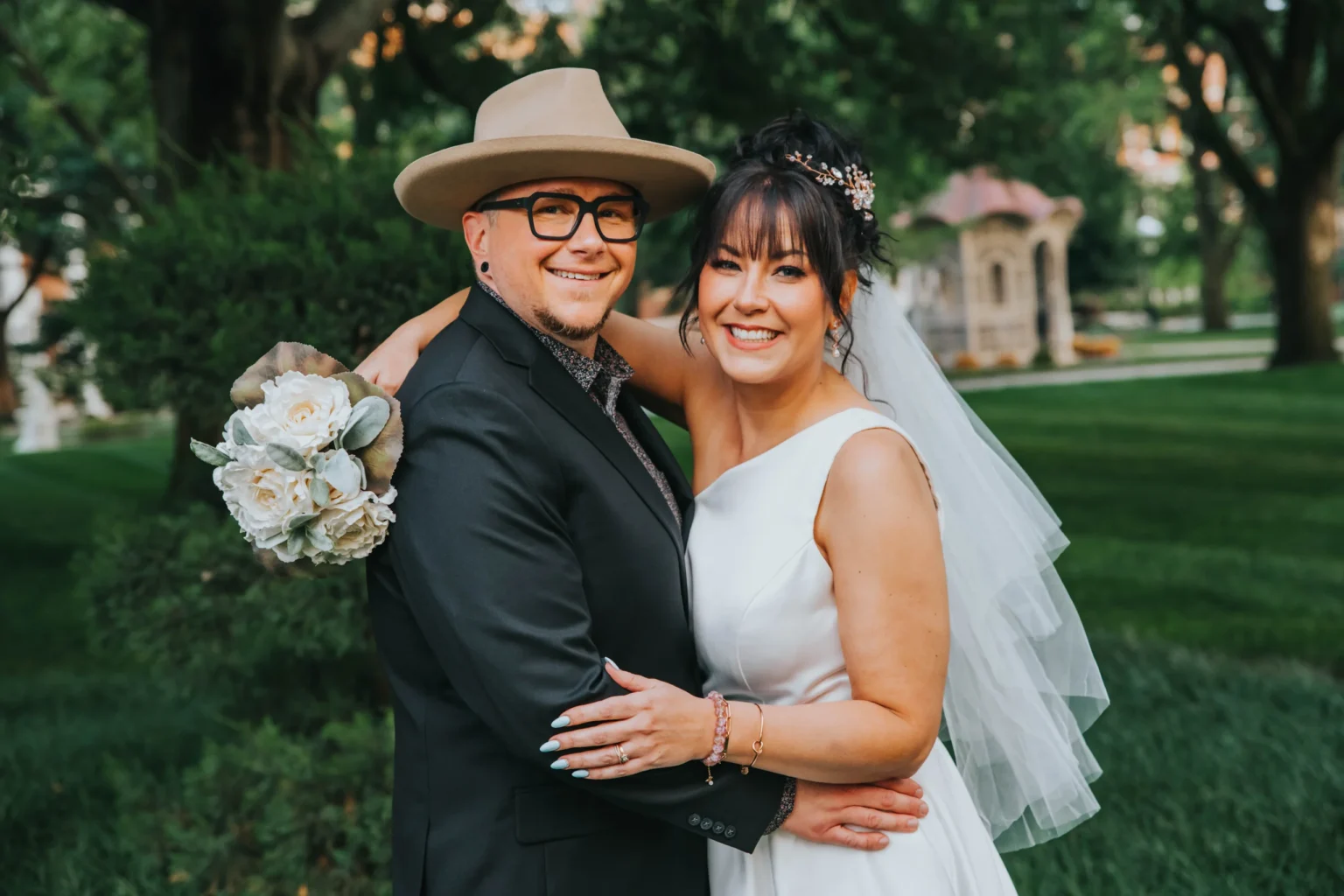 Elopement Wedding A smiling couple dressed in wedding attire pose outdoors. The person on the left wears a hat, glasses, and a black suit, while the person on the right is in a white bridal gown with a veil and a bouquet of white flowers. They embrace each other in a green, landscaped garden setting. Elopements Inc