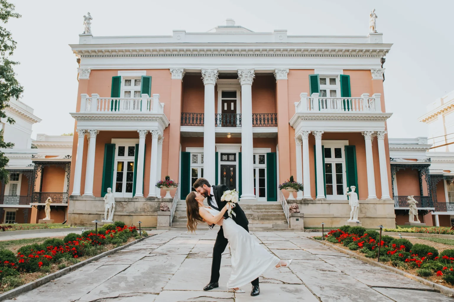 Elopement Wedding A couple dressed in wedding attire shares a romantic dance pose in front of a grand mansion with columns and green shutters. The groom dips the bride, who holds a bouquet, amidst a backdrop of manicured gardens and statues, creating a picturesque and elegant scene. Elopements Inc