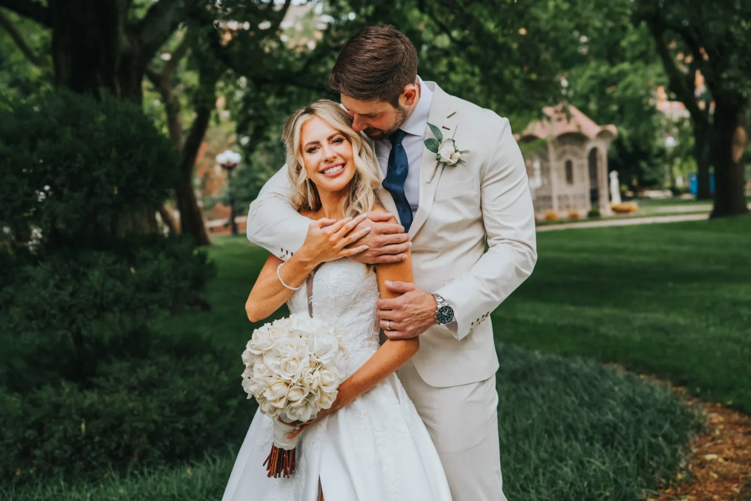 Elopement Wedding A bride in a white wedding dress holds a bouquet of white roses, smiling broadly. Behind her, a groom in a light-colored suit embraces her from behind, also smiling. They stand outdoors in a lush, green park with trees and a stone structure in the background. Elopements Inc