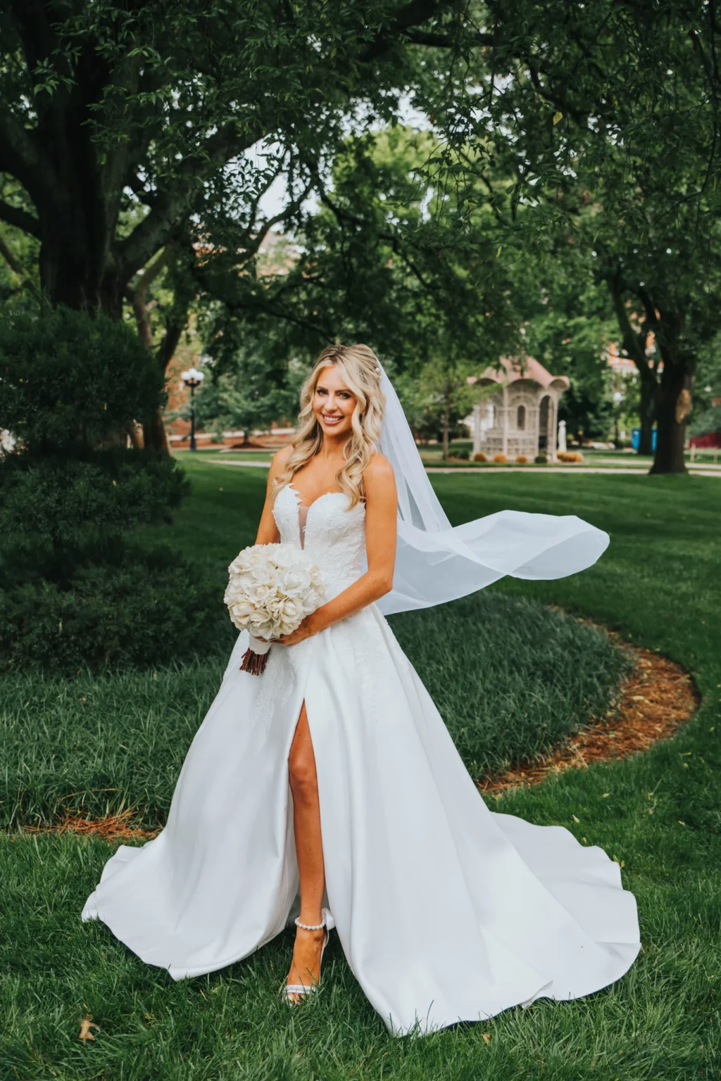 Elopement Wedding A bride stands outdoors in a lush green garden, wearing a strapless white wedding dress with a high slit and a long train. She holds a bouquet of white flowers and has a long veil blowing in the breeze. The background features a gazebo and several trees. She is smiling and looking at the camera. Elopements Inc