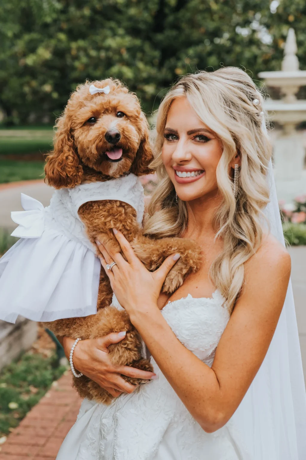 Elopement Wedding A bride with long, wavy blonde hair is smiling while holding a brown poodle dressed in a white dress. The bride, wearing a white wedding gown and veil, stands outside in a park. The background includes greenery and a white fountain. Both the bride and the dog look happy and well-groomed. Elopements Inc