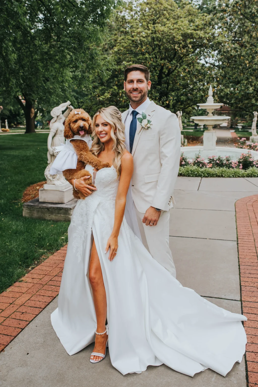 Elopement Wedding A smiling couple dressed in wedding attire poses outside. The bride is in a white strapless gown with a slit, while the groom wears a cream suit with a boutonnière. They hold a furry brown dog wrapped in a white cloth. They stand on a pathway with green trees and a fountain in the background. Elopements Inc