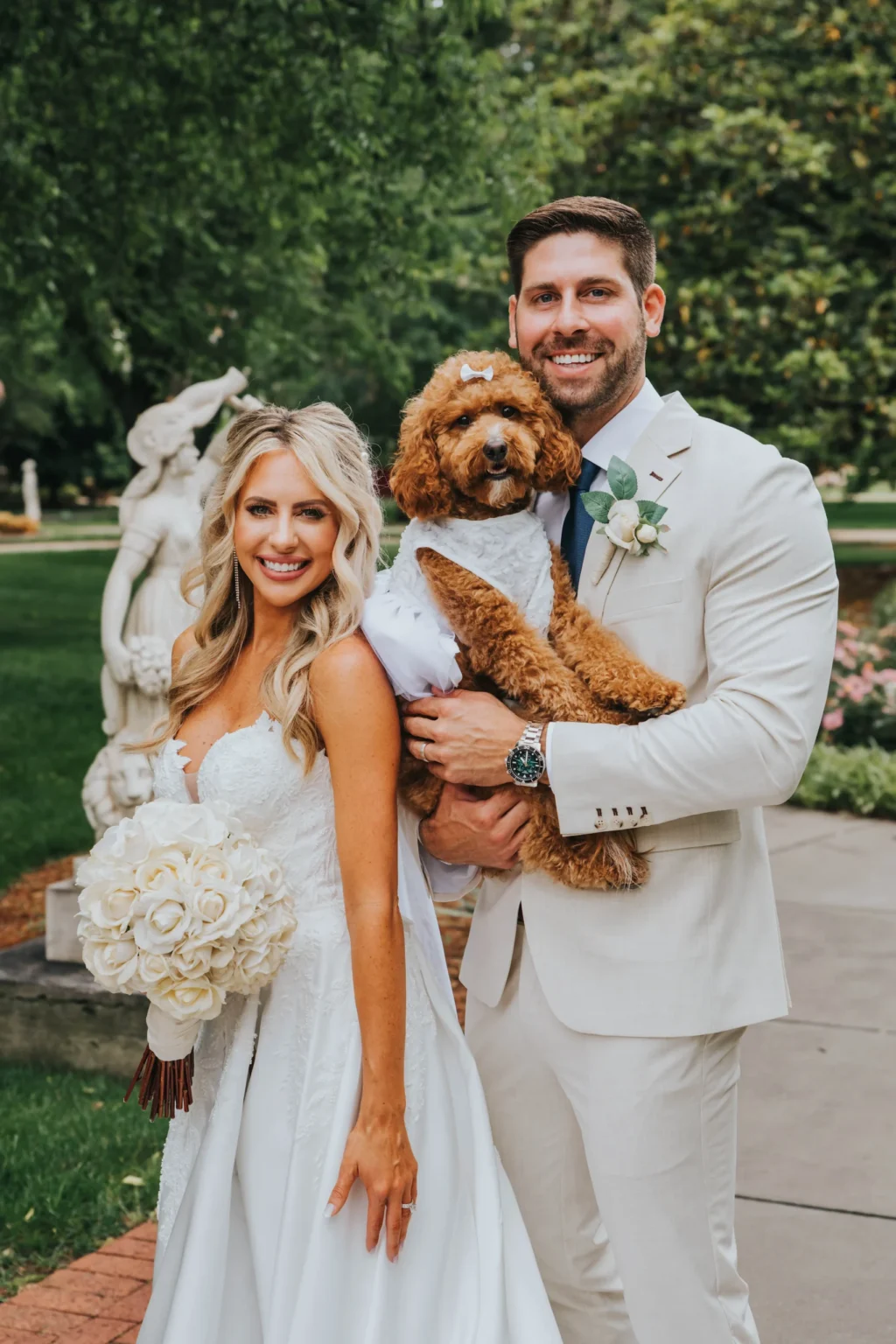 Elopement Wedding A bride and groom in elegant white attire smile while the groom holds a brown poodle adorned in a matching white dress. The bride holds a bouquet of white roses, and they stand in a lush garden with a stone sculpture behind them. The atmosphere is festive and joyful. Elopements Inc