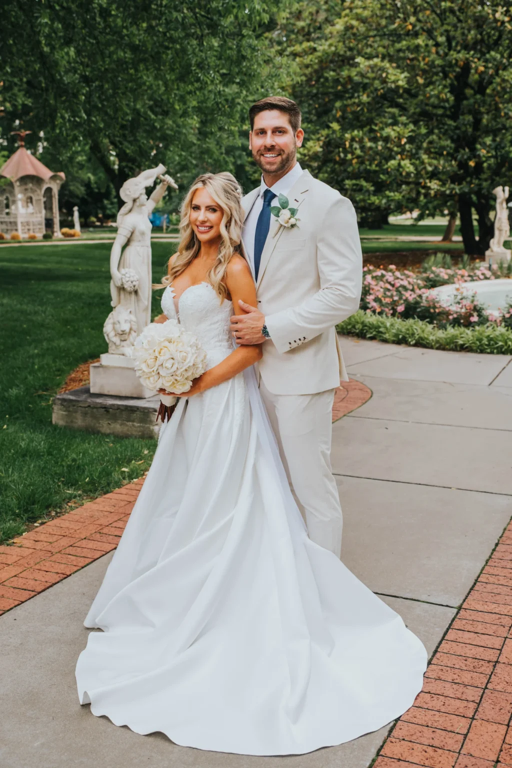 Elopement Wedding A bride and groom are posing outdoors on a brick and concrete path. The bride is wearing a white strapless gown, holding a bouquet of white flowers. The groom is wearing a white suit with a blue tie and boutonniere. They stand in front of greenery, garden statues, and a small gazebo. Elopements Inc