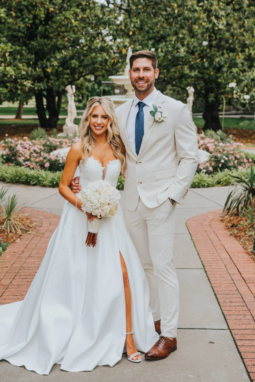 Elopement Wedding A bride dressed in a strapless white wedding gown holds a bouquet of white flowers while standing beside a groom in a light beige suit with a blue tie and white boutonniere. The couple is smiling and posing on a walkway in a garden with greenery and flowers in the background. Elopements Inc