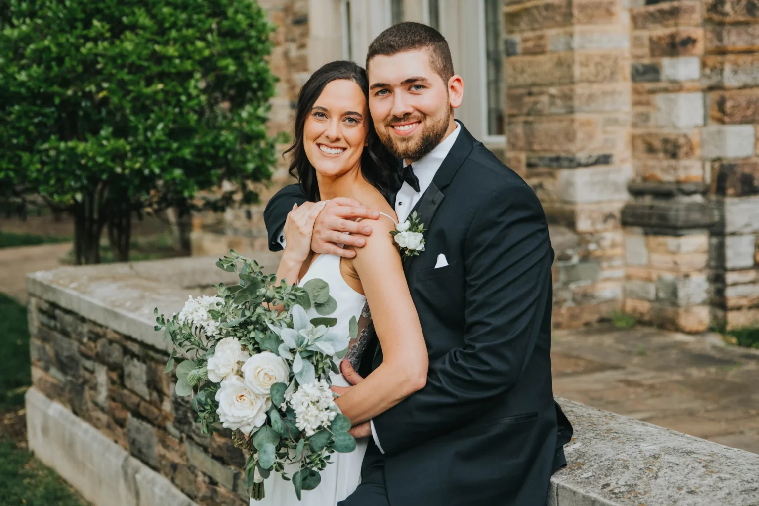 Elopement Wedding A smiling bride and groom pose together outdoors in front of a stone building. The bride, in a white dress, holds a bouquet of white roses and greenery. The groom, in a black tuxedo, embraces her from behind. Lush green foliage is visible in the background. Elopements Inc