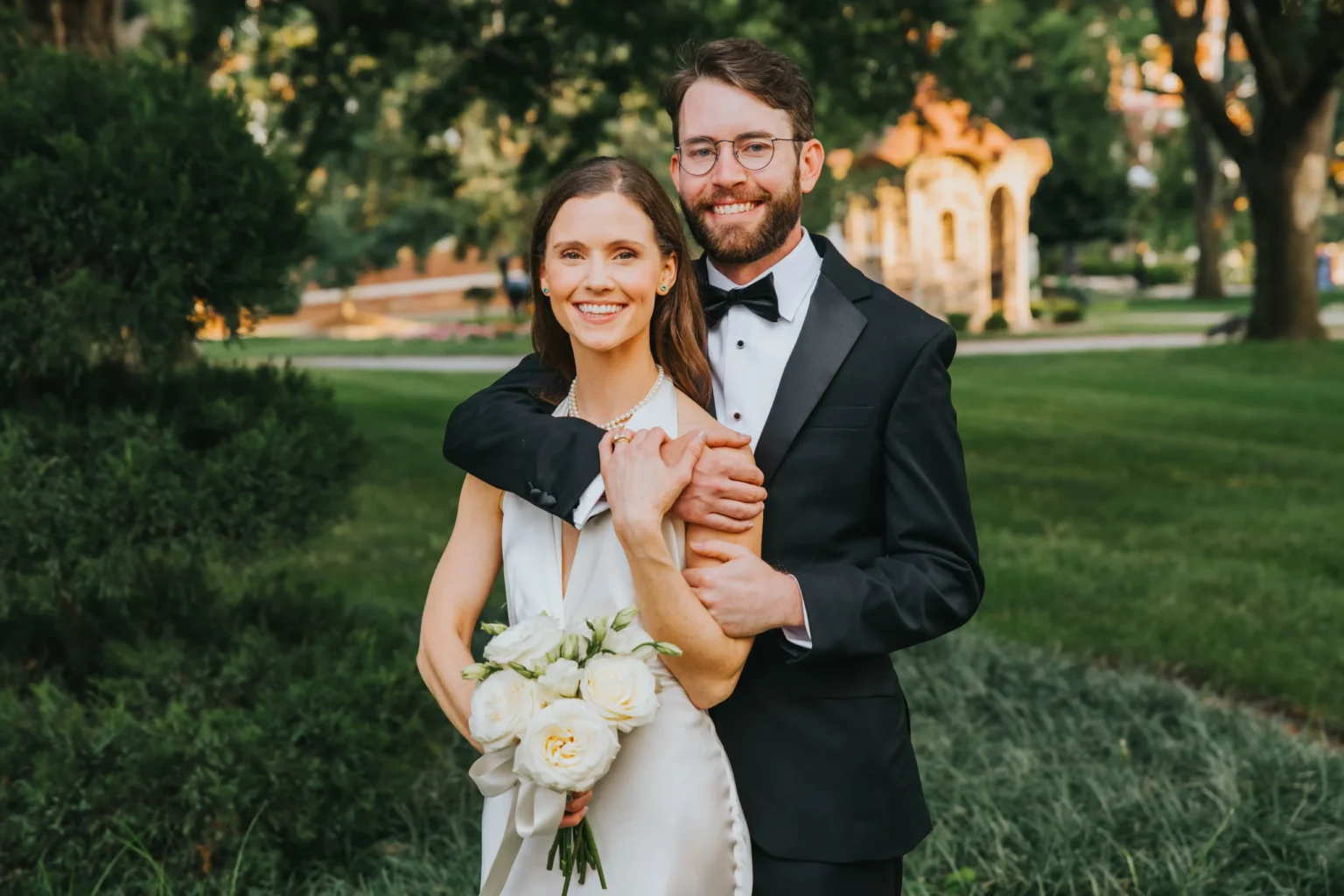 Elopement Wedding A happy couple poses outdoors on a lush green lawn. The woman, wearing a white sleeveless dress and holding a bouquet of white roses, stands in front of the man, who wears a black tuxedo and glasses. He wraps his arms around her shoulders. Both smile warmly at the camera. Elopements Inc