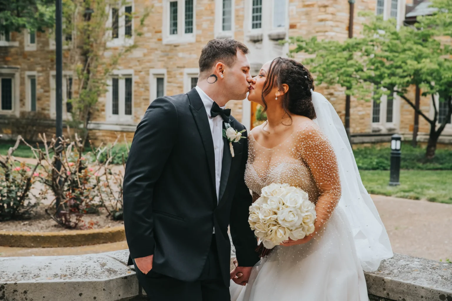 Elopement Wedding A couple dressed in wedding attire kiss outside. The groom wears a black tuxedo with a bow tie, while the bride wears a white gown with a veil and holds a bouquet of white roses. They are standing in front of a stone building with greenery and a courtyard pathway visible. Elopements Inc