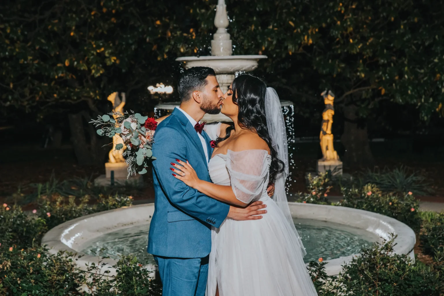 Elopement Wedding A bride and groom share a kiss in front of a fountain at night. The groom is wearing a blue suit, and the bride is in an off-the-shoulder white wedding dress with a long veil, holding a bouquet with red and white flowers. The background includes trees and garden lights. Elopements Inc