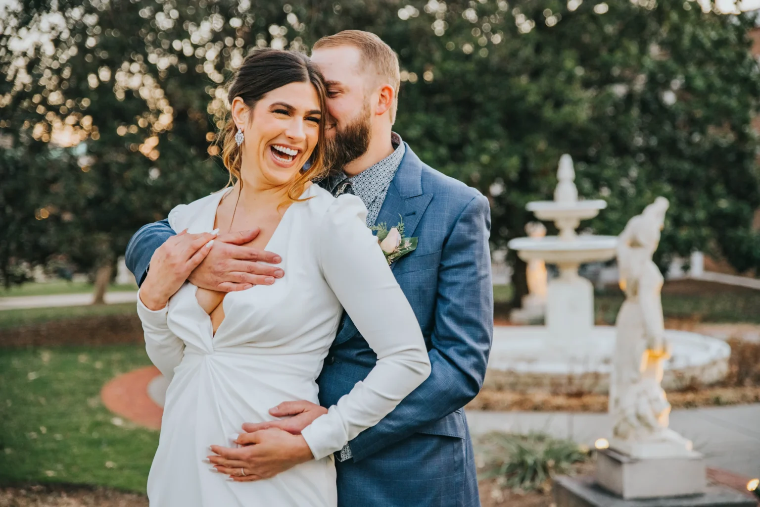 Elopement Wedding A couple stands in front of a fountain, sharing a joyful moment. The woman, wearing a white dress, smiles and leans back into the man, who embraces her from behind. The man wears a blue suit and has a beard. Both are laughing, framed by greenery in the background. Elopements Inc