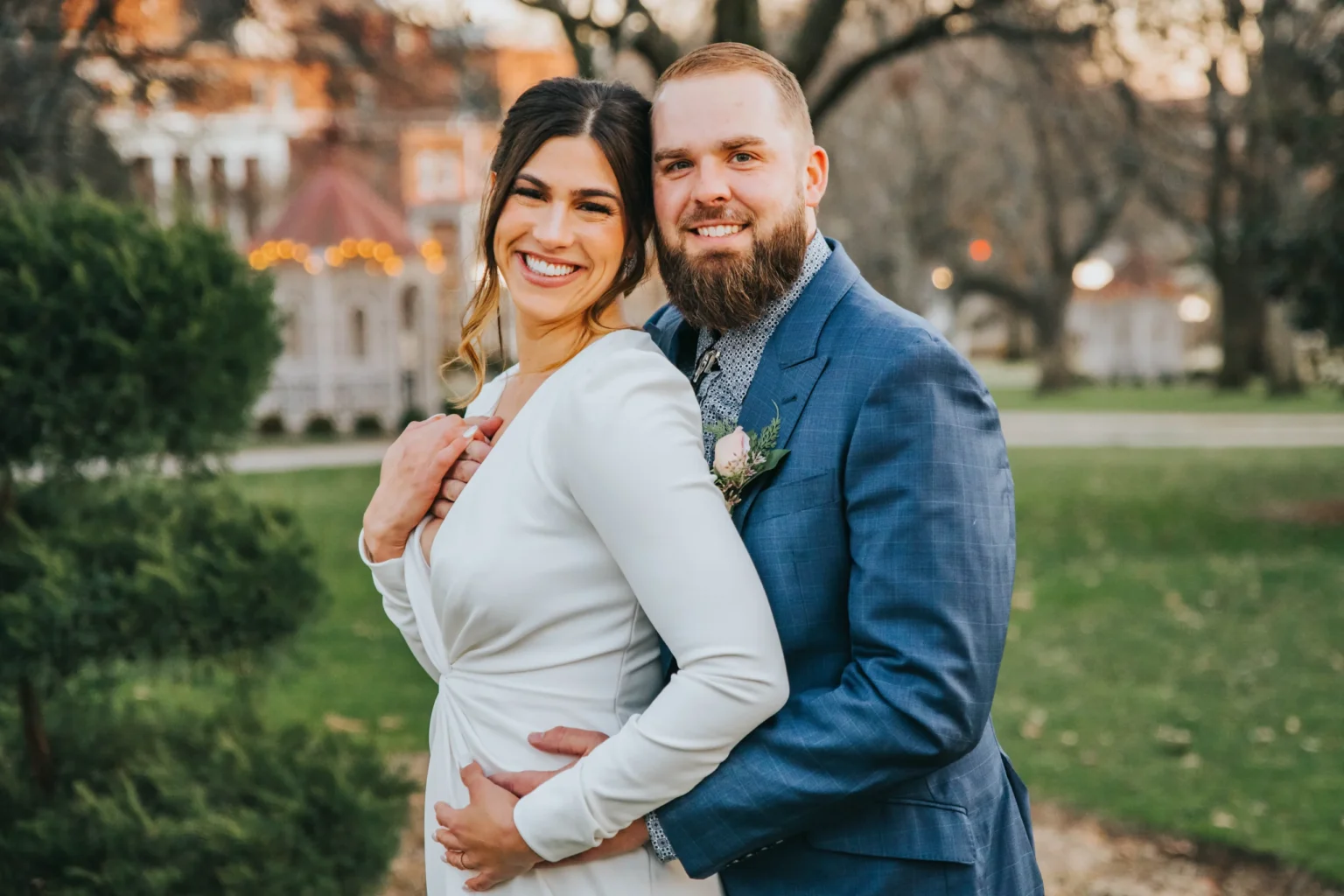 Elopement Wedding A smiling couple in formal attire poses outside on a green lawn with trees and a large building in the background. The woman, in a white dress, stands in front while the bearded man, in a blue suit, hugs her from behind with both hands around her waist. The scene is bright and cheerful. Elopements Inc