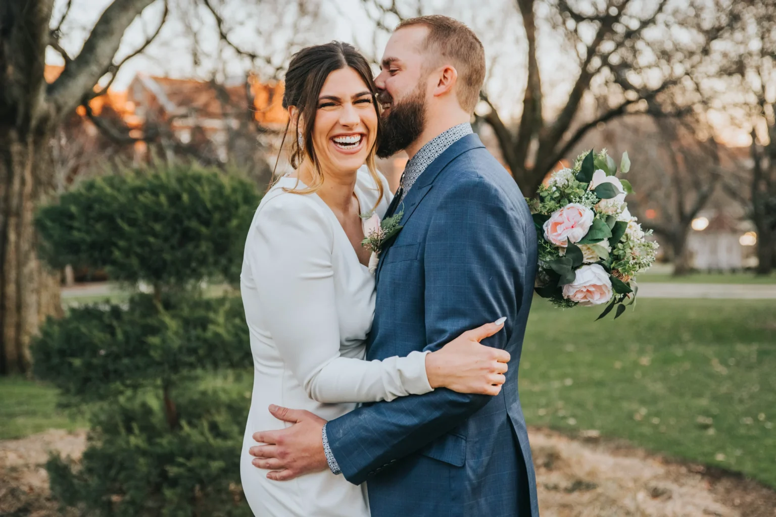 Elopement Wedding A bride and groom stand outdoors in a joyful embrace. The bride, in a white dress, is laughing. The groom, in a blue suit, is gently kissing her cheek. They are surrounded by trees, and the background shows a park setting with bare branches and a touch of sunlight. Elopements Inc