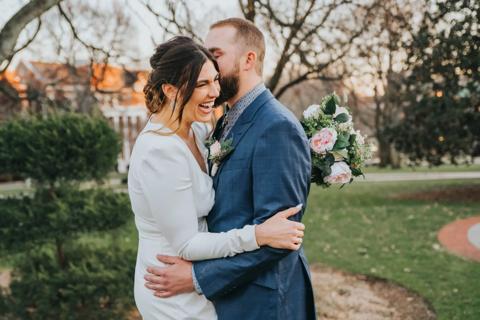 Elopement Wedding A joyous couple embraces in an outdoor setting. The woman, in a white dress, is smiling and laughing while the man, in a blue suit, gently kisses her on the cheek. The background features trees and a building with white columns, lit warmly by the setting sun. The man holds a bouquet of flowers. Elopements Inc