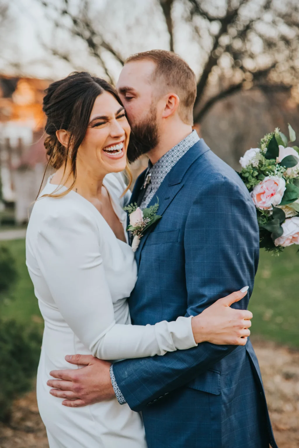 Elopement Wedding A joyful couple stands close in an outdoor setting. The woman, wearing a white dress, laughs heartily while the man, in a blue suit, grins and leans in as if whispering something to her. He holds a bouquet of flowers, featuring pink roses and greenery, in his hand. Trees and soft sunlight are in the background. Elopements Inc