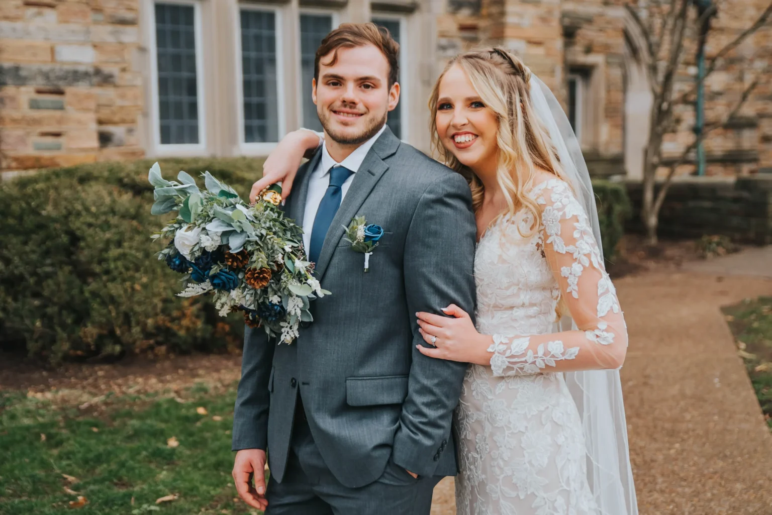 Elopement Wedding A bride and groom, smiling, stand outdoors in front of a stone building with large windows. The groom, in a gray suit and blue tie, has a blue boutonniere and the bride, in a lace gown with a veil, holds a bouquet of green, blue, and white flowers. The bride has her arm around the groom's shoulder. Elopements Inc