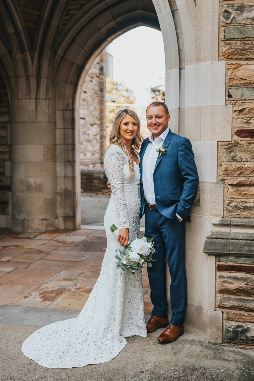 Elopement Wedding A bride and groom stand together outdoors under a stone archway. The bride is wearing a long-sleeved, lace wedding dress and holding a bouquet of white flowers. The groom is in a blue suit and white shirt. Both are smiling and leaning against a stone wall, with old stone buildings in the background. Elopements Inc