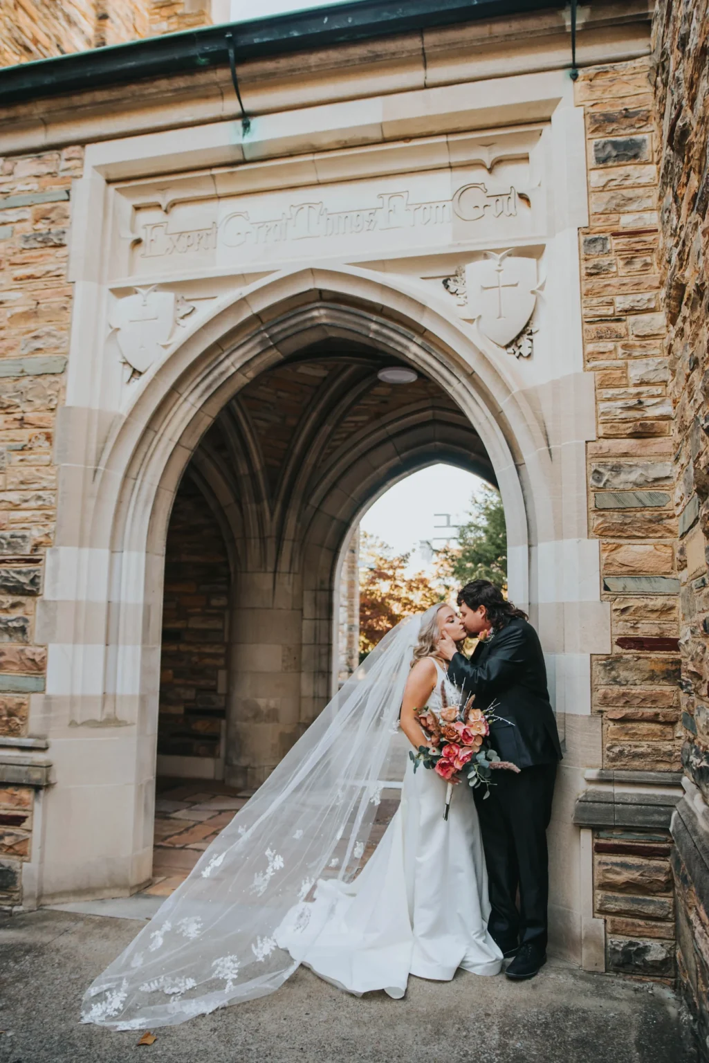 Elopement Wedding A newlywed couple shares a kiss under a stone archway decorated with carved inscriptions and symbols. The bride, in a long white dress with a flowing veil, holds a bouquet of pink flowers. The groom is dressed in a black suit and leans towards her. The background features textured stone walls. Elopements Inc