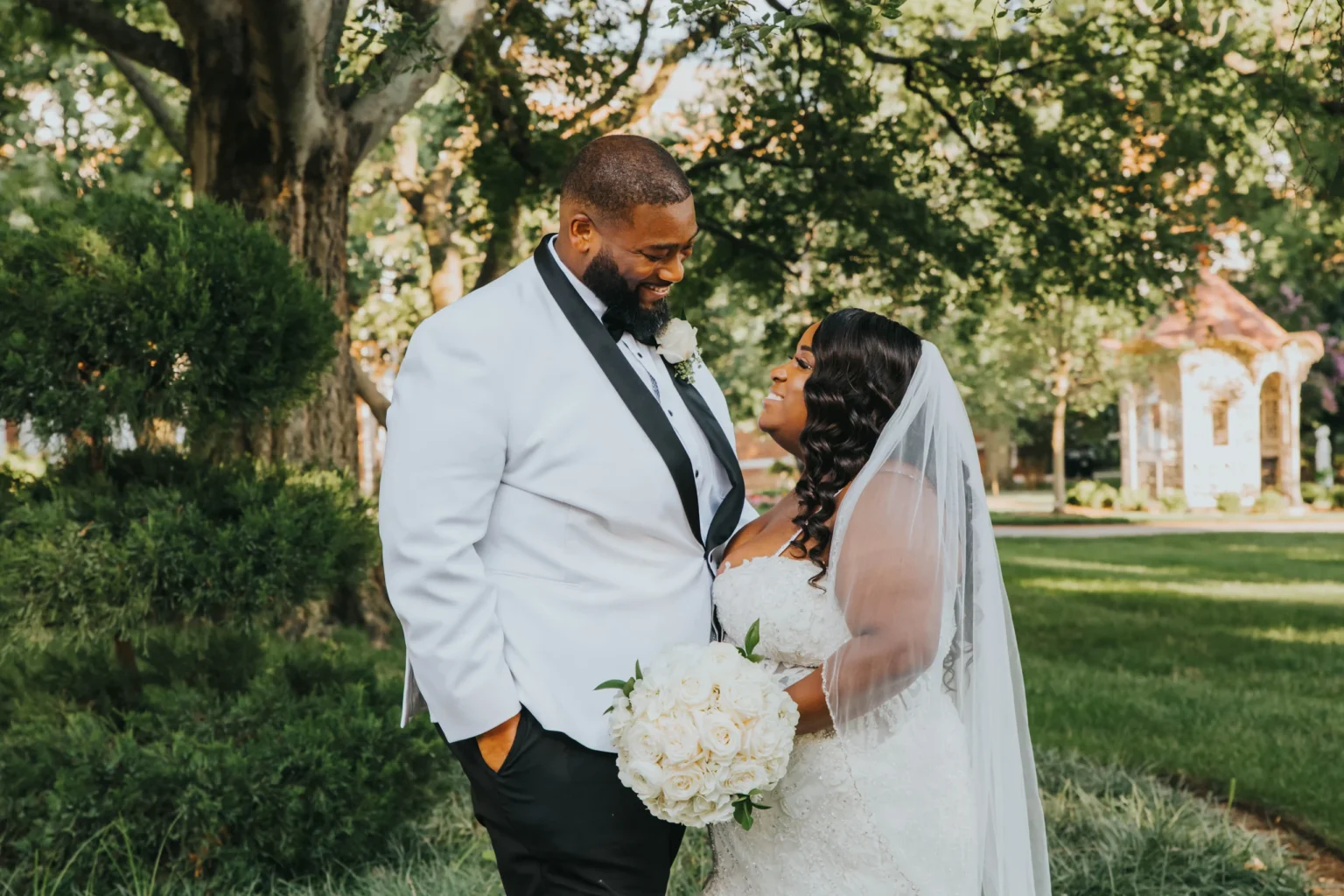 Elopement Wedding A newlywed couple stands close in a garden, smiling at each other. The groom wears a white tuxedo jacket with a black lapel, and the bride is in a strapless white wedding dress with a veil, holding a bouquet of white roses. The background includes trees, greenery, and a white gazebo. Elopements Inc