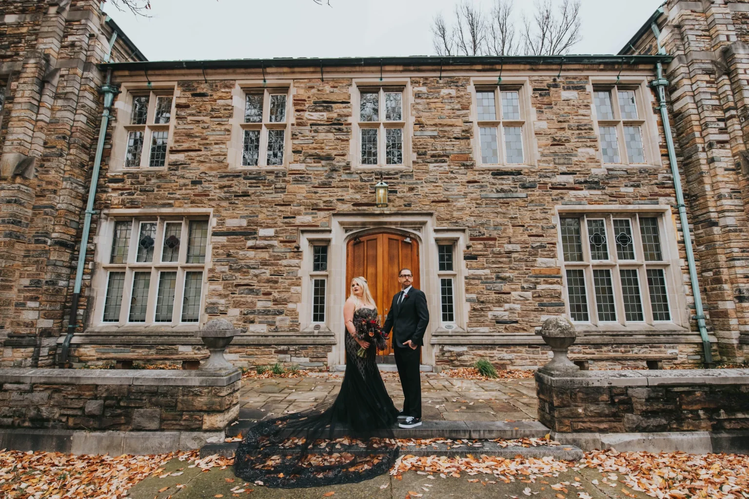 Elopement Wedding A couple stands in front of a large brick building with multiple windows. The woman in a black gown holds a bouquet and the man wears a suit. Both are holding hands and looking towards the camera. The ground is scattered with autumn leaves, and the scene has an elegant, timeless feel. Elopements Inc