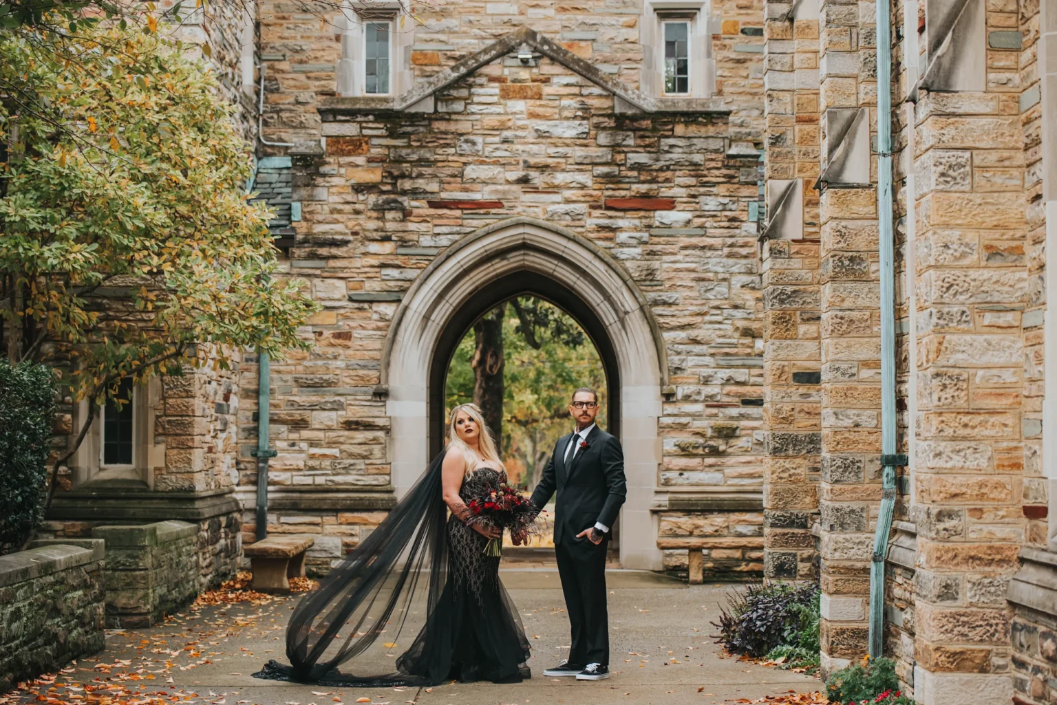 Elopement Wedding A couple stands in front of a historic stone building with a large archway. The woman is in a black gown with a long, flowing veil and holds a bouquet of red flowers. The man wears a black suit with a red tie and glasses. They are flanked by autumn foliage and fallen leaves on the pathway. Elopements Inc