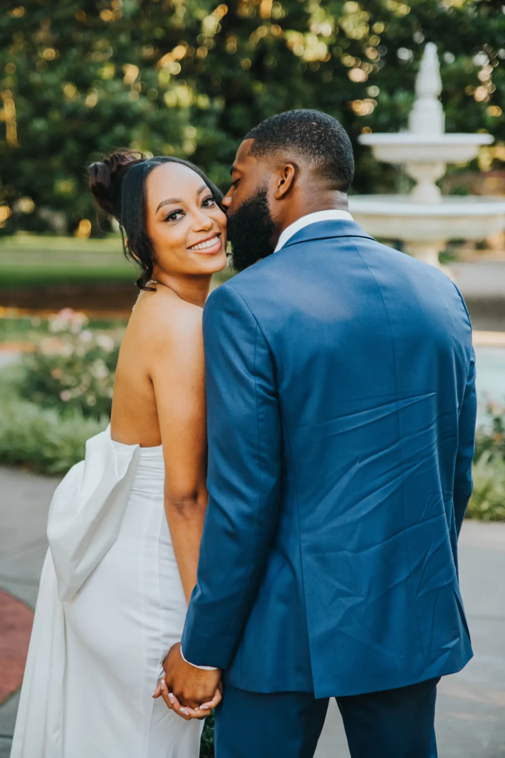 Elopement Wedding A bride in a strapless white dress smiles while holding hands with the groom, who is wearing a blue suit. They are outdoors with greenery and a white fountain blurred in the background. The groom leans in to kiss the bride's cheek as she looks at the camera. Elopements Inc