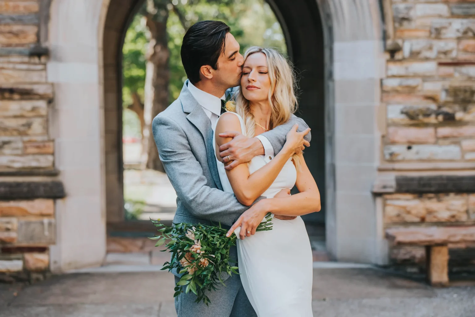 Elopement Wedding A couple stands embracing in front of a stone archway. The man, wearing a gray suit, kisses the woman's forehead as he holds her tightly. The woman, in a white dress, holds a bouquet of flowers and has her eyes closed. The background features a stone building and greenery. Elopements Inc