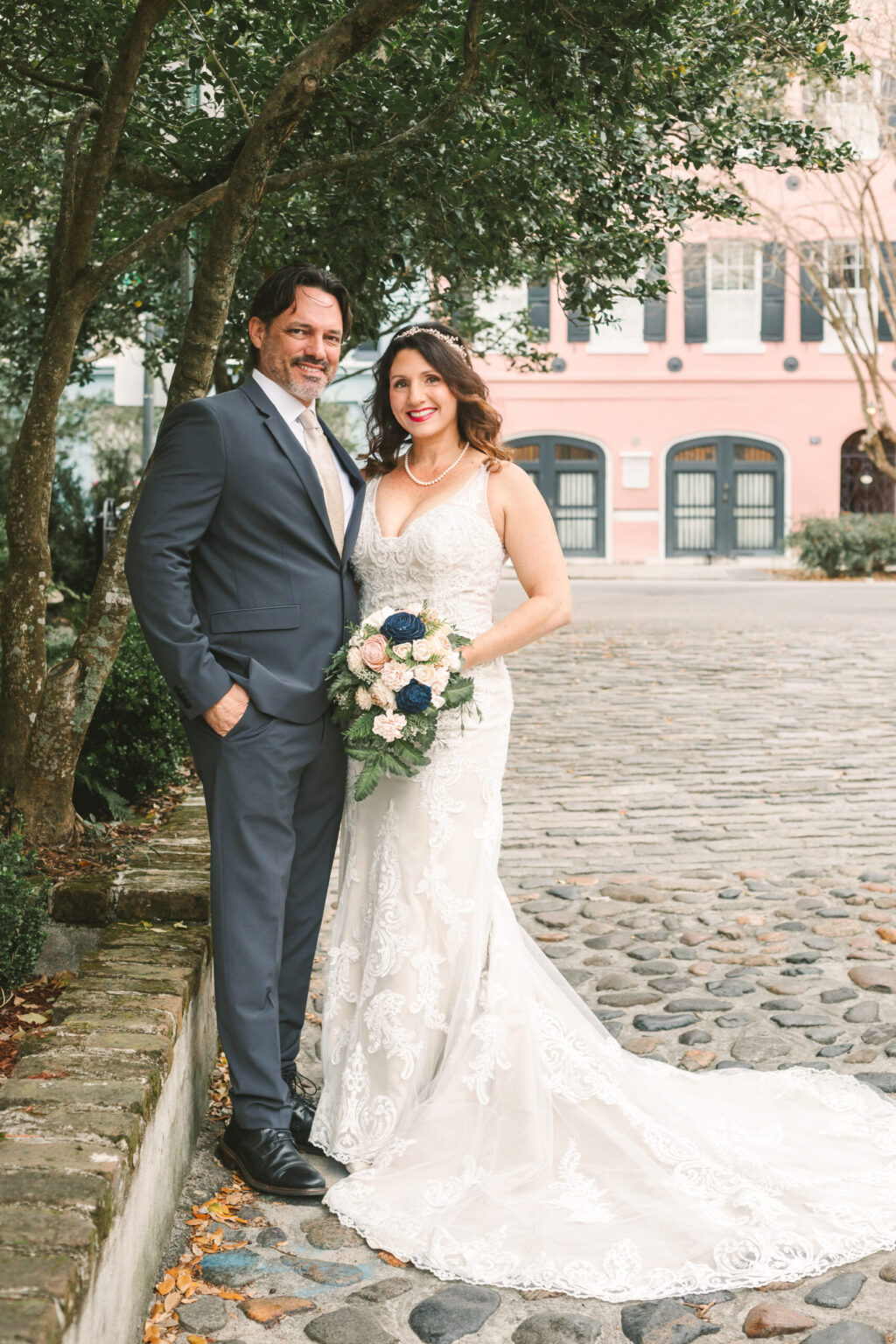 Elopement Wedding A bride and groom pose outdoors on a cobblestone street, capturing the essence of an intimate elopement. The bride wears a white lace gown and holds a bouquet of white and blue flowers. The groom is dressed in a grey suit with no tie. They stand under a tree, with buildings in pastel tones visible in the background. Elopements Inc