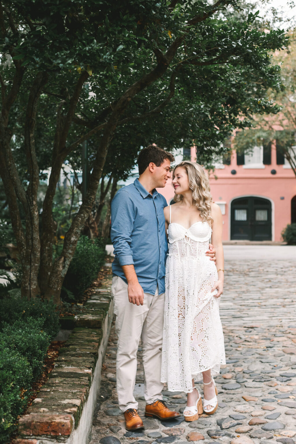 Elopement Wedding A couple, seemingly ready to elope, stands closely together on a cobblestone path, smiling at each other. The man wears a blue shirt and beige pants, while the woman wears a white eyelet dress. Trees surround them, and a pink building with large windows is visible in the background. Elopements Inc