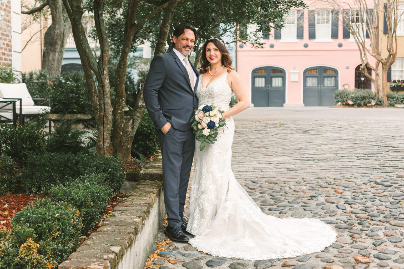 Elopement Wedding A couple stands smiling on a cobblestone street. The man wears a gray suit, and the woman, in a lace wedding dress, holds a bouquet. They are positioned near a tree-lined garden with a brick border. In the background are colorful buildings and garages, creating a picturesque setting perfect for an elopement. Elopements Inc