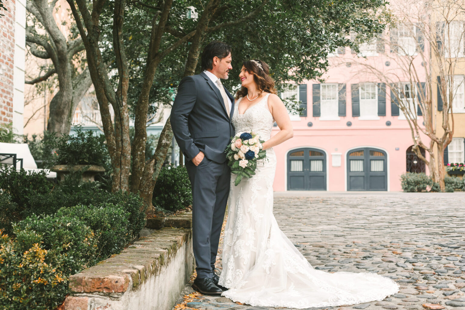 Elopement Wedding A bride and groom stand on a cobblestone path surrounded by greenery and historic buildings. The groom wears a dark suit and tie, while the bride, in an elegant white lace gown holding a bouquet of flowers, smiles at him. Their private elopement is filled with romance and joy as they gaze at each other. Elopements Inc