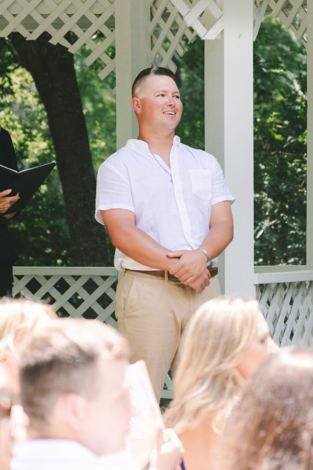 Elopement Wedding A person with short hair, wearing a white short-sleeve button-up shirt and beige pants, stands smiling under a white lattice gazebo. There are people with blurred faces in the foreground, possibly guests at an elopement, while the background features green trees and dappled sunlight. Elopements Inc