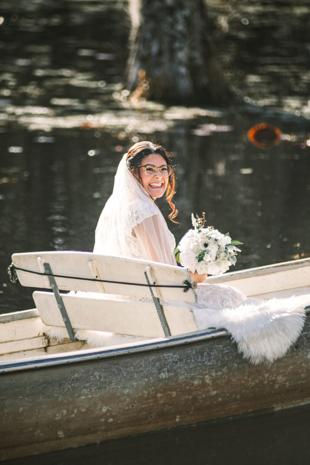 Elopement Wedding A bride in a white dress and veil, holding a bouquet of white flowers, smiles while sitting in a small wooden rowboat. The boat is on a calm body of water, perfect for their intimate elopement. She sits on a fur-covered cushion as the scene is bathed in warm, natural light. Elopements Inc