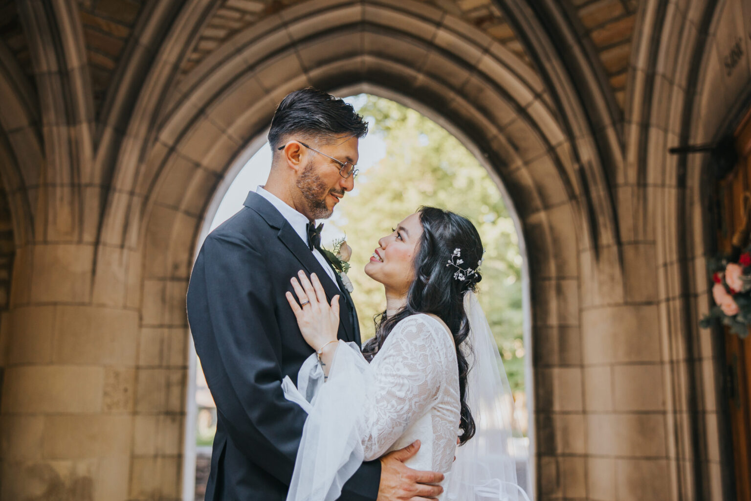 Elopement Wedding A couple dressed in wedding attire stand closely together under a stone archway. The groom, in a dark suit and glasses, holds the bride, who is in a white lace gown with floral hair adornments. They gaze lovingly into each other's eyes, with sunlight filtering through the archway behind them, capturing the essence of their intimate elopement. Elopements Inc