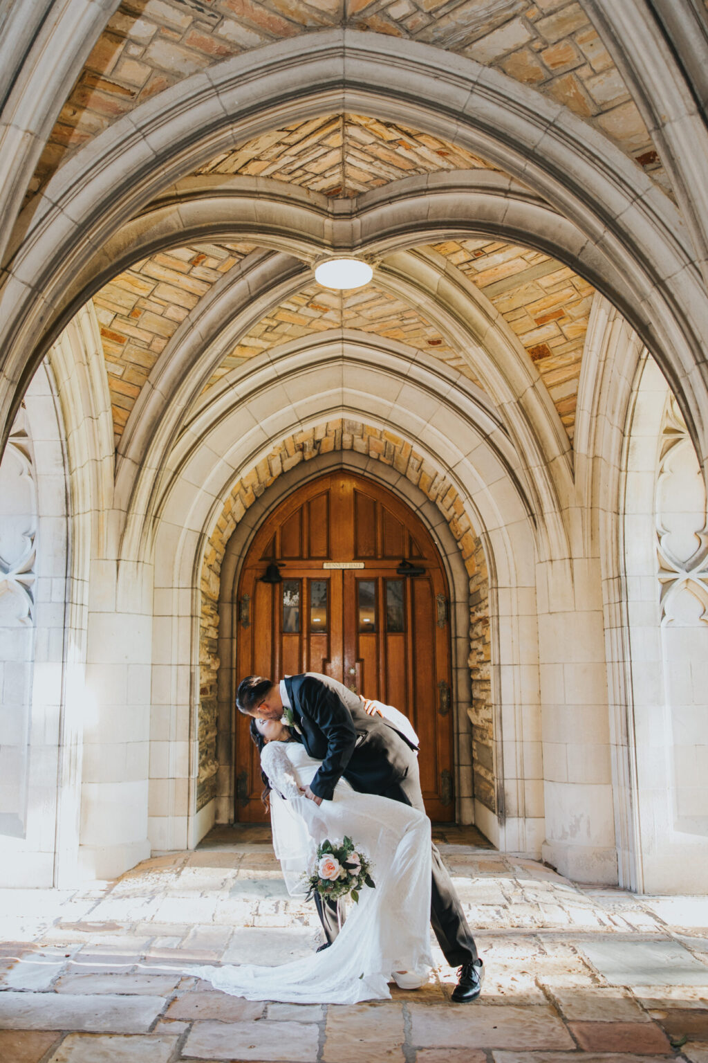 Elopement Wedding A newlywed couple shares a kiss as the groom dips the bride under a stone archway. The bride holds a bouquet of flowers, and both are dressed in formal wedding attire. The intricate archway, perfect for elopements, leads to a wooden door with a stone path beneath them. Elopements Inc