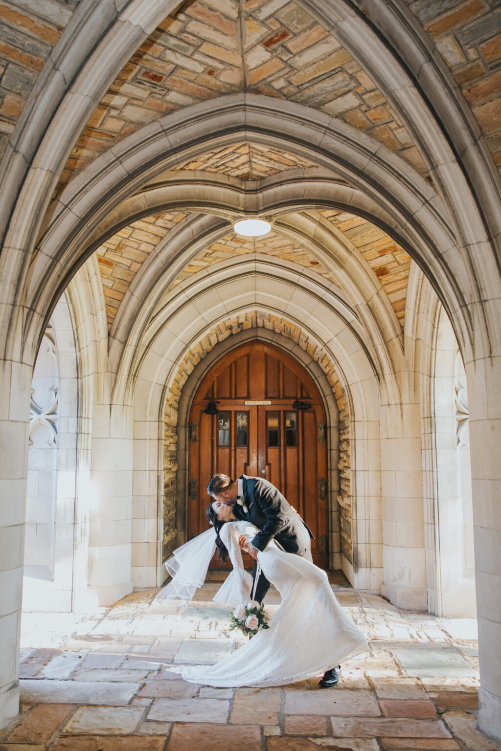 Elopement Wedding A newly married couple shares a romantic kiss under a stone archway. The groom, dressed in a black suit, dips the bride who wears a flowing white gown and holds a bouquet. Sunlight filters through the arches, illuminating the scene and the wooden door in the background—a perfect elopement moment. Elopements Inc