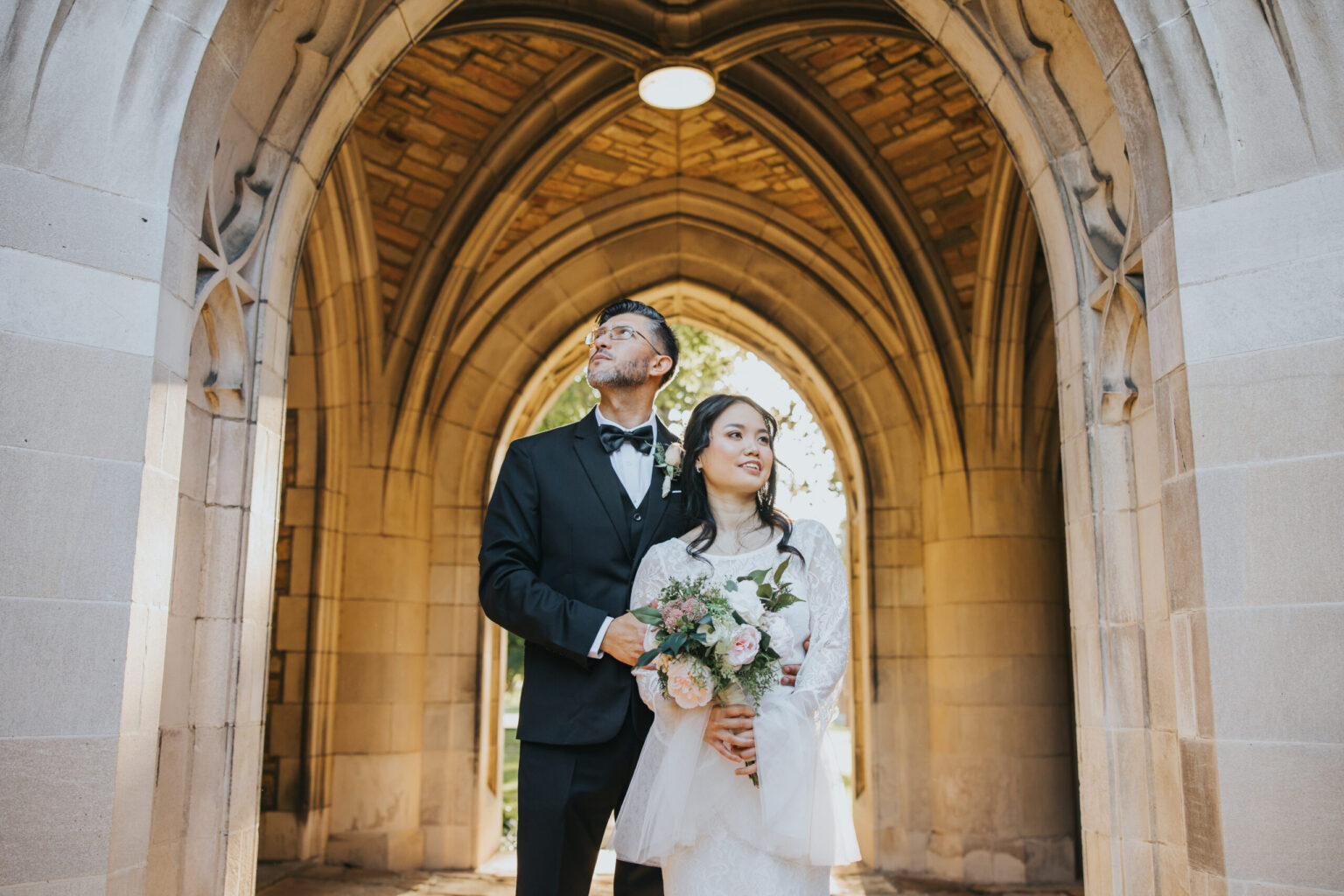 Elopement Wedding A bride and groom stand under a stone archway. The groom wears a black suit and bow tie, while the bride wears a white lace wedding dress and holds a bouquet of flowers. Both, having chosen to elope, look off into the distance with serene expressions, framed by the elegant architectural details above them. Elopements Inc