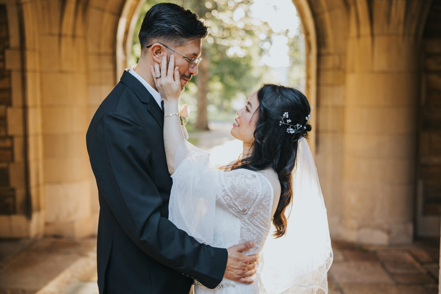 Elopement Wedding A bride and groom stand closely under a stone archway, eloping in an intimate ceremony. The bride gently touches the groom's face. He wears glasses and a dark suit, while she wears a white lace dress and a veil with small flowers. Sunlight filters through trees in the background, creating a warm, romantic atmosphere. Elopements Inc