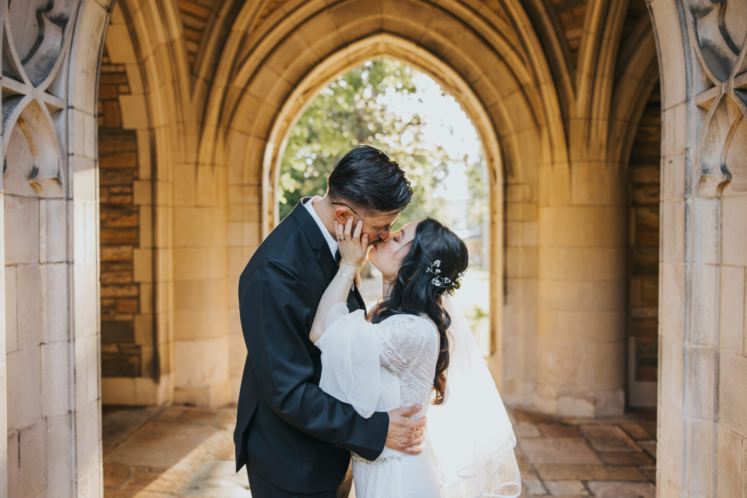 Elopement Wedding A couple, dressed in formal attire, shares a kiss under a stone archway. The groom is in a black suit and the bride wears a white wedding dress with a veil and floral hair accessories. Sunlight illuminates them through the arch, creating a warm and romantic atmosphere perfect for elopements. Elopements Inc