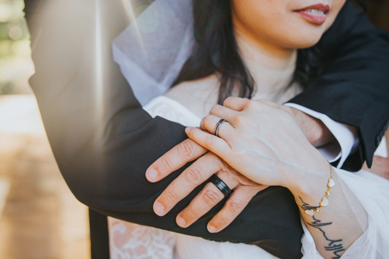 Elopement Wedding A close-up of a bride and groom from the shoulders down, captured during their elopement. The groom, in a black suit, embraces the tattooed bride from behind. Both display wedding rings—hers is delicate while his is thicker. Sun rays filter through, creating a warm, intimate atmosphere. Elopements Inc