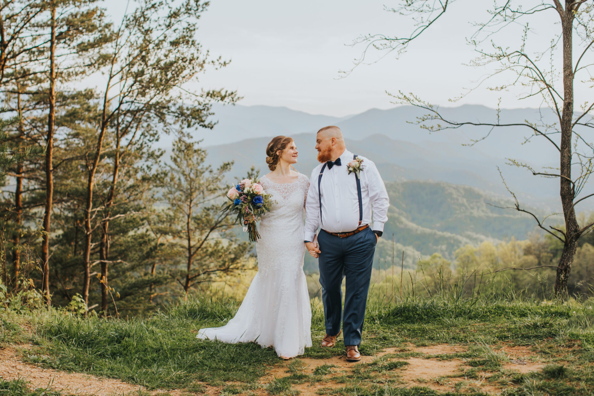 Elopement Wedding A bride and groom stand hand-in-hand, smiling at each other, against a backdrop of rolling hills and forested mountains. The bride wears a long white lace dress and holds a bouquet of flowers. The groom dons a white shirt with a bow tie and blue suspenders. This intimate elopement captures the lush and serene scenery perfectly. Elopements Inc