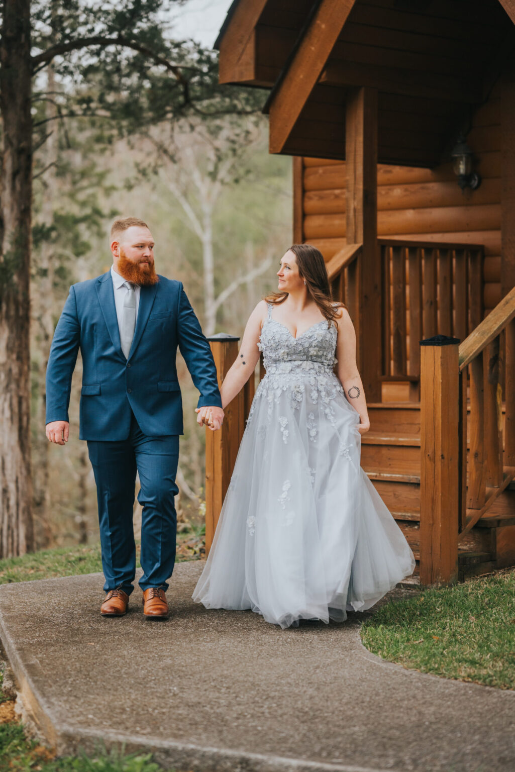 Elopement Wedding A couple walks hand in hand down a path in front of a wooden cabin, embodying the spirit of elopement. The man wears a blue suit with a white shirt and brown shoes. The woman wears a light gray, sleeveless gown with floral appliqués and a tulle skirt. They both look at each other, smiling as trees frame the backdrop. Elopements Inc