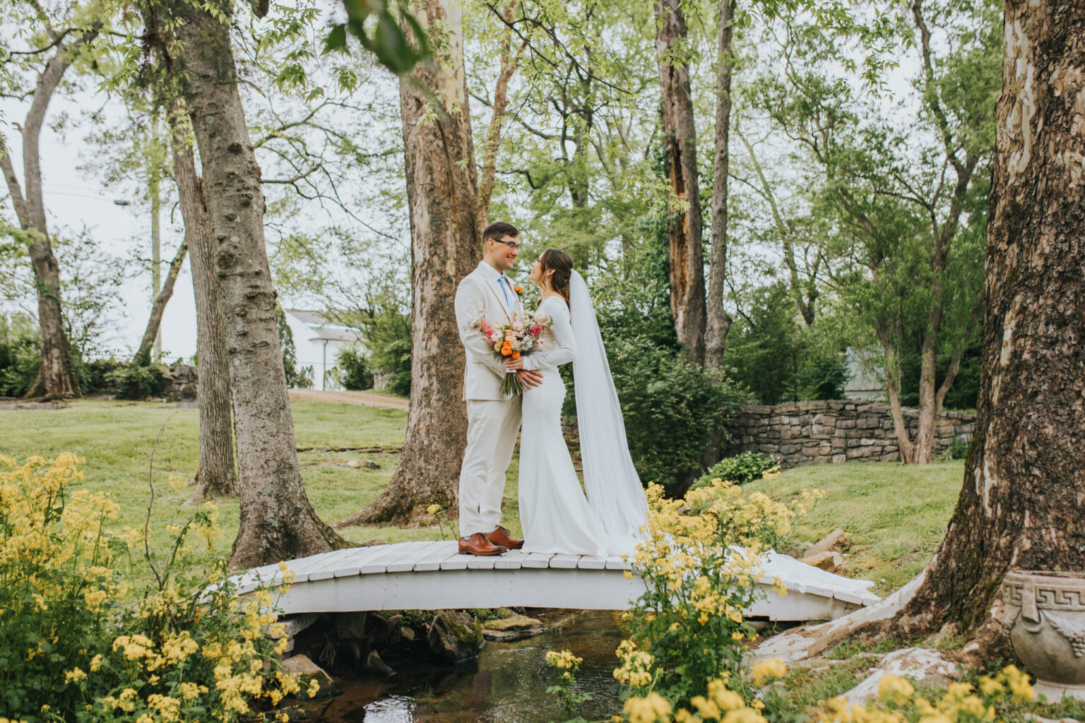 Elopement Wedding A bride and groom stand together on a small white bridge over a narrow stream in a lush, green woodland area, eloping to their hidden paradise. The bride wears a flowing white dress and veil, holding a bouquet of colorful flowers. The groom is in a light tan suit and smiles at the bride as they stand close amidst vibrant yellow flowers. Elopements Inc